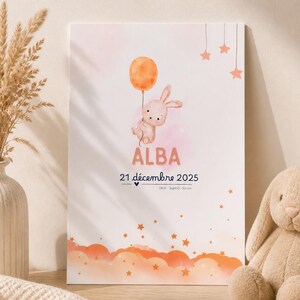 Peut inclure: Impression encadrée avec une illustration à l'aquarelle d'un lapin tenant un ballon orange. L'impression comprend le nom "ALBA" et la date "21 décembre 2025". Le bas de l'impression présente des nuages oranges et des étoiles.