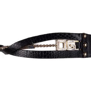 Może przedstawiać: Czarny skórzany pasek z złotym łańcuszkiem i dużą złotą klamrą z literami "DG" w białych kryształach.