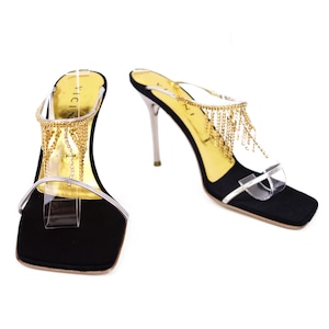 May include: Black high-heeled sandals with gold chain fringe and clear straps. The heels are silver and the soles are black. The sandals have a square toe and a gold insole.