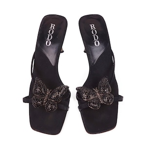 Op de afbeelding: Een paar zwarte sandalen met hak en vlinderversiering. De sandalen hebben een vierkante neus en een dun enkelbandje. De vlinder is gemaakt van donkere strass-steentjes. De merknaam "RODO" staat op de binnenzool.