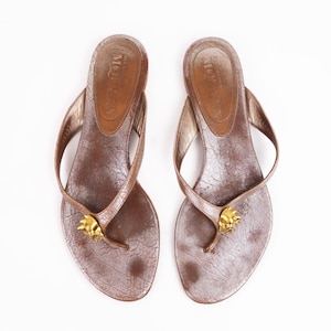 Könnte beinhalten: Ein Paar braune Ledersandalen mit Riemen und goldenen Akzenten. Die Sandalen haben ein schlichtes Design und eine bequeme Passform.