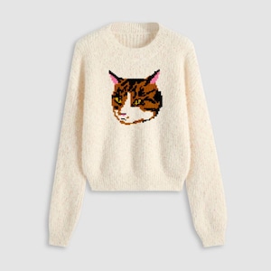 May include: Cream-colored sweater featuring a pixelated cat face design. The cat's face is brown, black, white, and yellow, with pink ears. The sweater has a crew neck and long sleeves, perfect for a cozy and casual look.