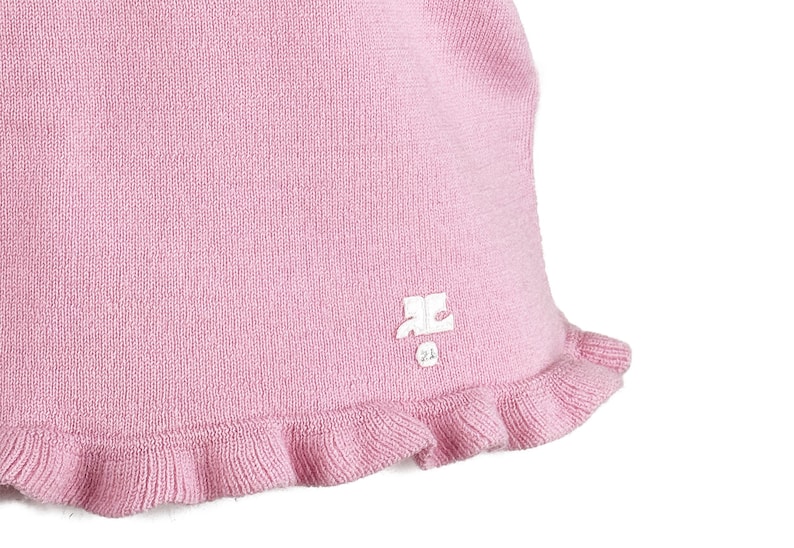 Op de afbeelding: Lichtroze gebreid kledingstuk met een gerimpelde rand. De stof heeft een getextureerde uitstraling. Een klein wit logo is zichtbaar bij de rand. Het kledingstuk lijkt een hoed of een soortgelijke hoofdbedekking te zijn.