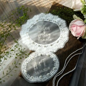 May include: Two white, oval wedding favours with intricate lace edging and pearl embellishments. The top favour reads "Stefan & Jenny" and "Oct. 21, 2026." The bottom favour reads "A & T" and "18.10.2026."