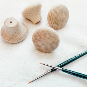 Könnte beinhalten: Vier unfertige Holzknöpfe und zwei Pinsel auf einer weißen, flauschigen Oberfläche.