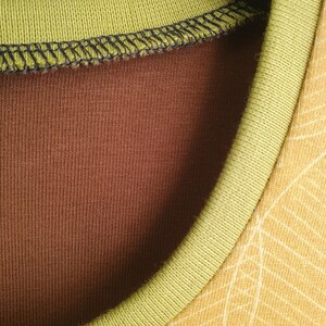 Op de afbeelding: Een shirt met ronde hals met een effen bruine binnenstof en een mosterdgele buitenstof met een wit lijnpatroon. De halslijn is afgewerkt met een geribbelde olijfgroene boord en zwarte stiksels langs de binnenste kraagnaad.