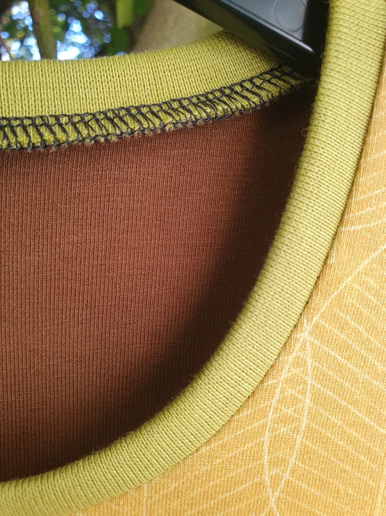 Op de afbeelding: Een shirt met ronde hals met een effen bruine binnenstof en een mosterdgele buitenstof met een wit lijnpatroon. De halslijn is afgewerkt met een geribbelde olijfgroene boord en zwarte stiksels langs de binnenste kraagnaad.