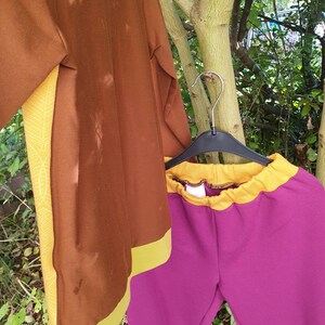 Op de afbeelding: Een bruine top met lange mouwen met gele zijpanelen met een wit bladpatroon en een gele zoom, gecombineerd met een magentakleurige broek met een gele elastische tailleband, beide hangend aan een zwarte plastic hanger aan een boomtak.