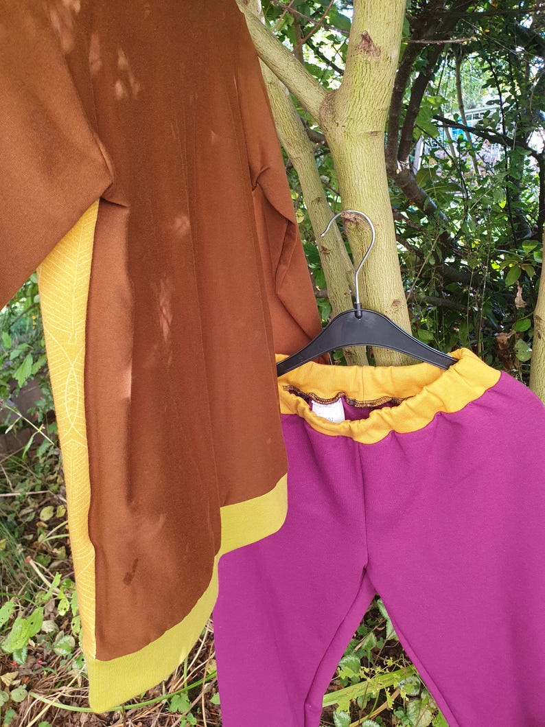 Op de afbeelding: Een bruine top met lange mouwen met gele zijpanelen met een wit bladpatroon en een gele zoom, gecombineerd met een magentakleurige broek met een gele elastische tailleband, beide hangend aan een zwarte plastic hanger aan een boomtak.