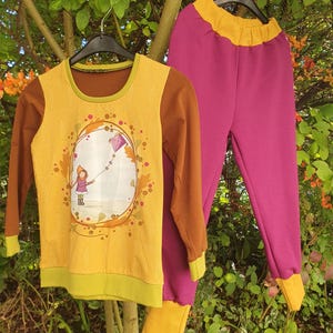 Op de afbeelding: Een set bestaande uit een shirt met lange mouwen en een broek. Het shirt heeft een geel voorpand met een afbeelding van een persoon die een paarse vlieger oplaat, bruine mouwen en groene boorden. De broek is magenta met een gele tailleband.