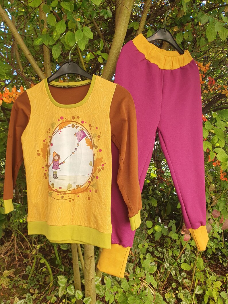 Op de afbeelding: Een set bestaande uit een shirt met lange mouwen en een broek. Het shirt heeft een geel voorpand met een afbeelding van een persoon die een paarse vlieger oplaat, bruine mouwen en groene boorden. De broek is magenta met een gele tailleband.