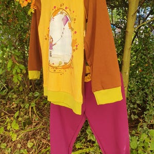 Op de afbeelding: Een set bestaande uit een shirt met lange mouwen en een broek. Het shirt heeft bruine mouwen, een gele voorkant met een subtiel bladpatroon en een afbeelding van een persoon die een paarse vlieger oplaat. De broek is effen magenta van kleur.