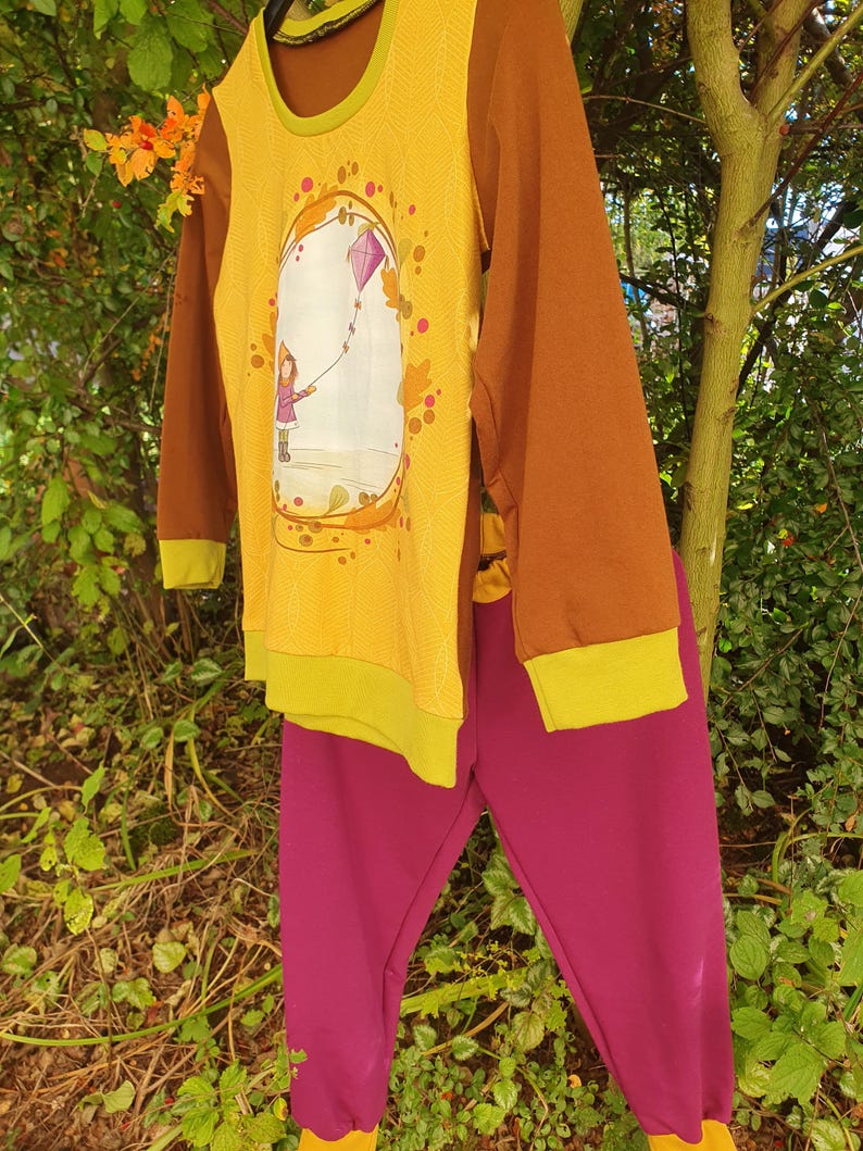 Op de afbeelding: Een set bestaande uit een shirt met lange mouwen en een broek. Het shirt heeft bruine mouwen, een gele voorkant met een subtiel bladpatroon en een afbeelding van een persoon die een paarse vlieger oplaat. De broek is effen magenta van kleur.