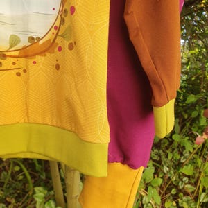 Op de afbeelding: Een sweatshirt met lange mouwen met een mosterdgeel voorpand met een wit bladpatroon en een cirkelvormige afbeelding, een effen donker magenta zijpaneel, een bruine mouw, een mosterdgele manchet en een limoengroene zoom.