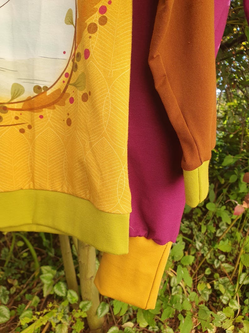 Op de afbeelding: Een sweatshirt met lange mouwen met een mosterdgeel voorpand met een wit bladpatroon en een cirkelvormige afbeelding, een effen donker magenta zijpaneel, een bruine mouw, een mosterdgele manchet en een limoengroene zoom.