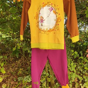 Op de afbeelding: Een set bestaande uit een shirt met lange mouwen en een broek. Het gele shirt met bruine mouwen heeft een afbeelding van een persoon die een paarse vlieger oplaat. De broek is effen magenta en heeft gele boorden aan de onderkant van de pijpen.