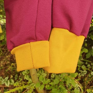 Op de afbeelding: Een kledingstuk met lange mouwen in diep magenta stof met contrasterende geribbelde boorden in mosterdgeel. De mouwen zijn bij de pols geplooid waar ze samenkomen met de effen gele boorden, wat een gladde, gebreide textuur laat zien.