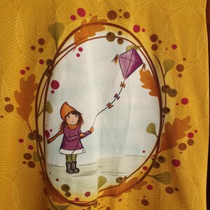 Op de afbeelding: Een mosterdgeel shirt met een groene kraag en een ovale print van een kind in een paarse jas en oranje muts dat een paarse vlieger oplaat, omringd door herfstbladeren, eikels en kleine rode cirkels.