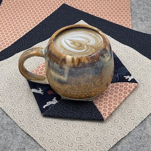 May include: A brown ceramic mug with a heart-shaped design in the foam, on a patchwork coaster. The coaster has geometric patterns in cream, navy, and coral, with a grey felt base. A cosy coffee or tea setting.