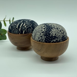 May include: Two dome-shaped pin cushions set into small, round, natural-toned wooden bowls. The left cushion features a dark blue fabric with a white floral pattern, while the right cushion has a dark blue fabric with a white lace-like design.