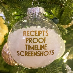 Könnte beinhalten: Ein transparenter Weihnachtsbaumschmuck aus Glas mit einem weißen, flauschigen Innenleben. Der Schmuck ist mit goldenen Buchstaben beschriftet: "RECEIPTS PROOF TIMELINE SCREENSHOTS".
