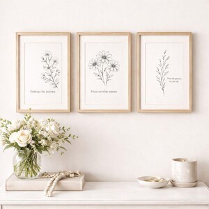 May include: Three framed botanical art prints with line drawings of flowers and foliage. Each print has a light wood frame and a white background. The prints have inspirational text: "Embrace the journey," "Focus on what matters," and "Trust the process of your life."