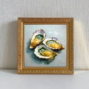 May include: A framed oil painting featuring three open oysters. The oysters have a creamy yellow interior and are set against a light blue background. The frame is ornate and gold-colored.
