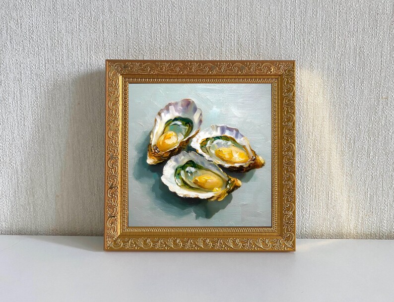 May include: A framed oil painting featuring three open oysters. The oysters have a creamy yellow interior and are set against a light blue background. The frame is ornate and gold-colored.