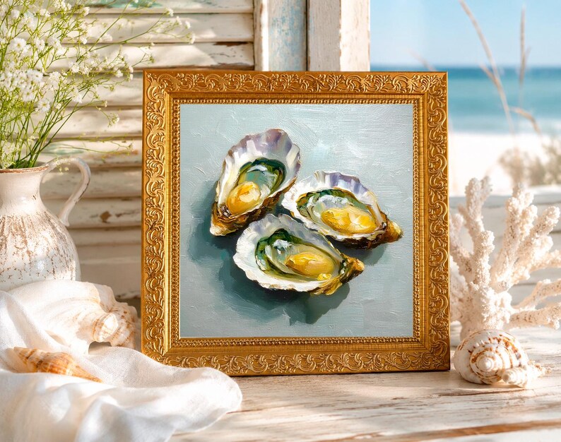 May include: A framed painting of three open oysters with yellow and green interiors, set against a light blue background. The painting is in a gold ornate frame, with a coastal theme. The scene includes a vase of white flowers and seashells.