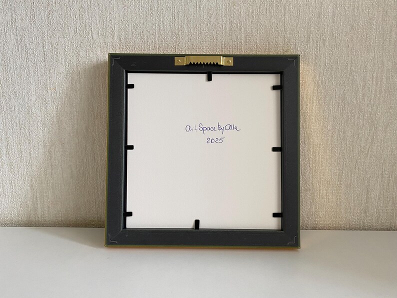 May include: Square framed artwork, viewed from the back. The frame is black with a gold-colored edge. The white backing has handwritten text that reads "Art + Space by Allie 2025". A sawtooth hanger is attached.