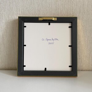May include: Back of a framed artwork with a black frame and a gold-colored edge. The white backing has the handwritten text "Art + Space by Allie 2025". The frame is square and has a sawtooth hanger.
