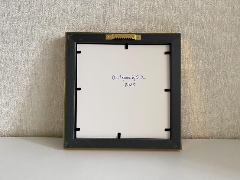 May include: Back of a framed artwork with a black frame and a gold-colored edge. The white backing has the handwritten text "Art + Space by Allie 2025". The frame is square and has a sawtooth hanger.