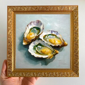 May include: An oil painting of three oysters in their shells, with a golden frame. The oysters have a yellow and green interior, set against a light blue background. The frame is ornate with a gold finish.