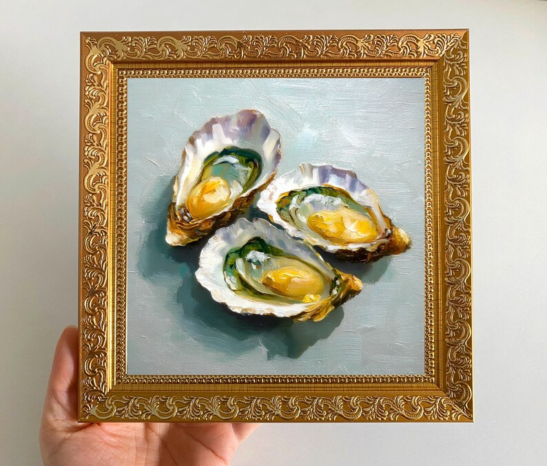 May include: An oil painting of three oysters in their shells, with a golden frame. The oysters have a yellow and green interior, set against a light blue background. The frame is ornate with a gold finish.