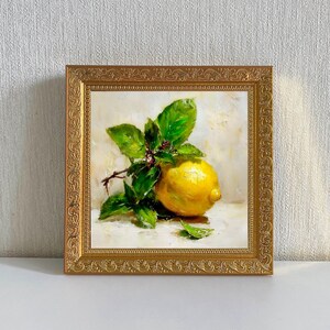 May include: A framed oil painting of a lemon with green leaves and small red berries. The lemon is yellow and sits on a white surface. The painting is in a gold ornate frame.