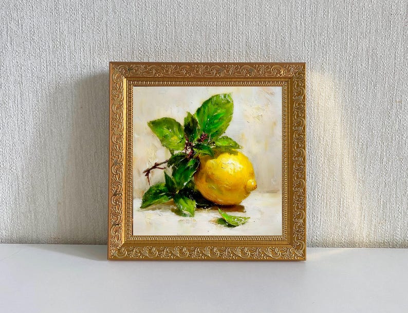 May include: A framed oil painting of a lemon with green leaves and small red berries. The lemon is yellow and sits on a white surface. The painting is in a gold ornate frame.