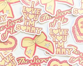 May include: Assortment of pink and yellow stickers with various designs, including hearts, bows, and text. The text on the stickers includes "I love you I'm sorry", "The Secret of Us", and "The Secret of Us".