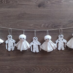 May include: A decorative garland featuring seven hanging ornaments suspended from a white string. The ornaments alternate between white fabric ghost figures with black button eyes and white fabric mummy figures wrapped in strips of white cloth.