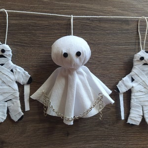 May include: A hanging decoration featuring two fabric mummy figures wrapped in white strips with black circular eyes and a central ghost figure made of white fabric with black eyes and a silver-toned metal chain draped across the front.