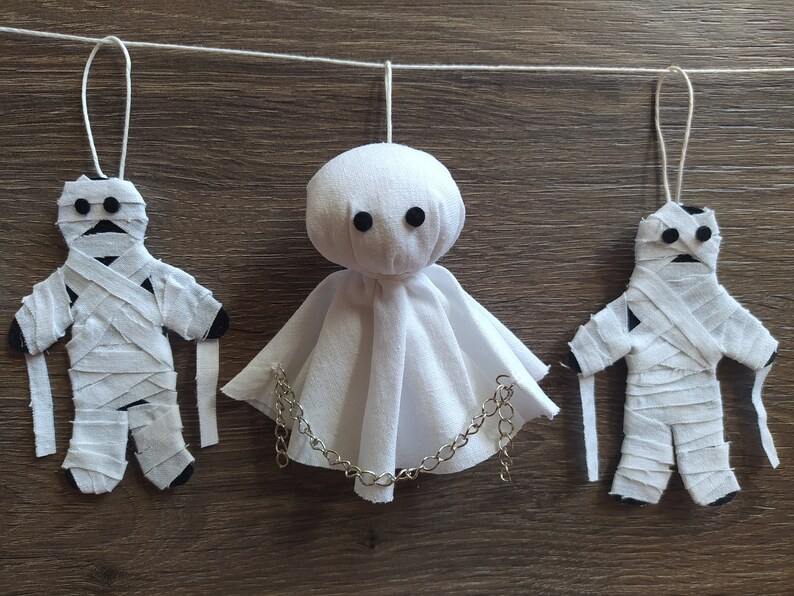 May include: A hanging decoration featuring two fabric mummy figures wrapped in white strips with black circular eyes and a central ghost figure made of white fabric with black eyes and a silver-toned metal chain draped across the front.