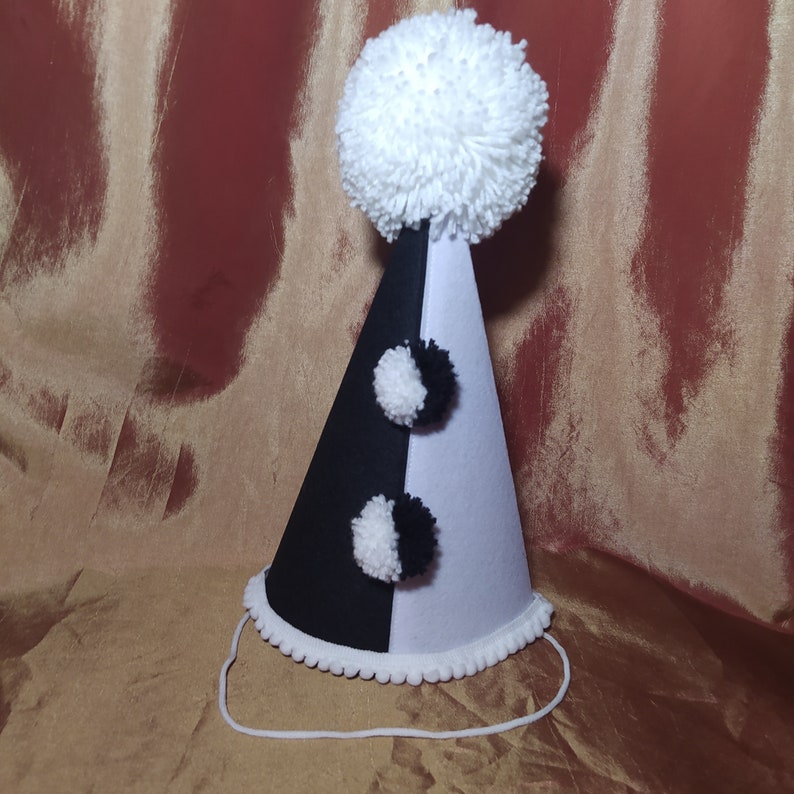 May include: A party hat with a black and white color scheme. The hat is cone-shaped, with a large white pom-pom on top and two smaller pom-poms on the front. A white string is attached for securing the hat.