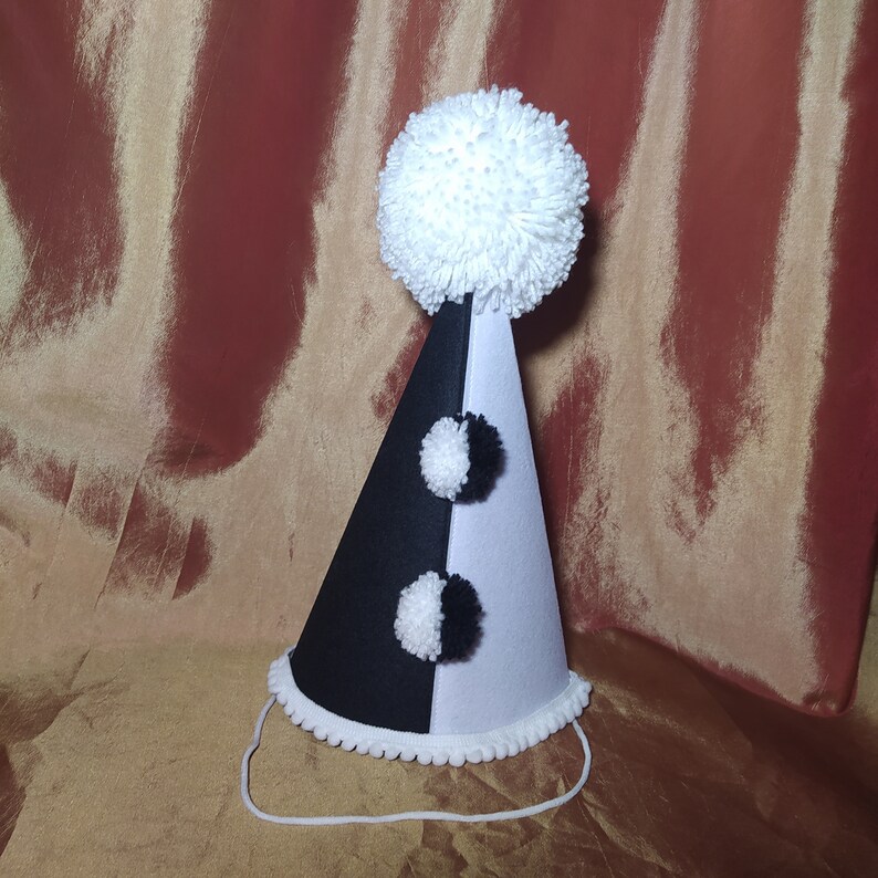May include: A black and white party hat with a large white pom-pom on top and two smaller pom-poms on the front. The hat has a white trim and a string for securing it.