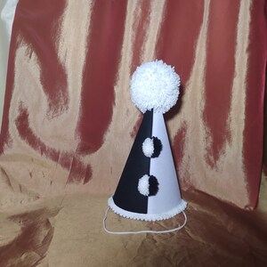 May include: A black and white party hat with a large white pom-pom on top. The cone-shaped hat has a black and white alternating pattern with two smaller white pom-poms. A white string is attached for securing the hat.