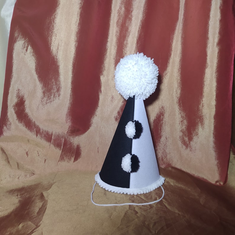 May include: A black and white party hat with a large white pom-pom on top. The cone-shaped hat has a black and white alternating pattern with two smaller white pom-poms. A white string is attached for securing the hat.