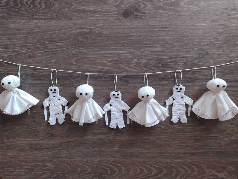 May include: A decorative garland featuring seven hanging ornaments suspended from a white string. The ornaments alternate between white fabric ghost figures with black button eyes and white fabric mummy figures wrapped in strips of white cloth.