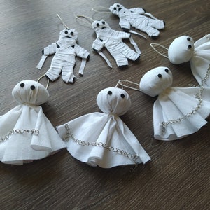 May include: Three white fabric ghost ornaments with black bead eyes and silver-toned chains draped across their skirts, alongside three white fabric mummy ornaments wrapped in strips with black bead eyes, all featuring small hanging loops.