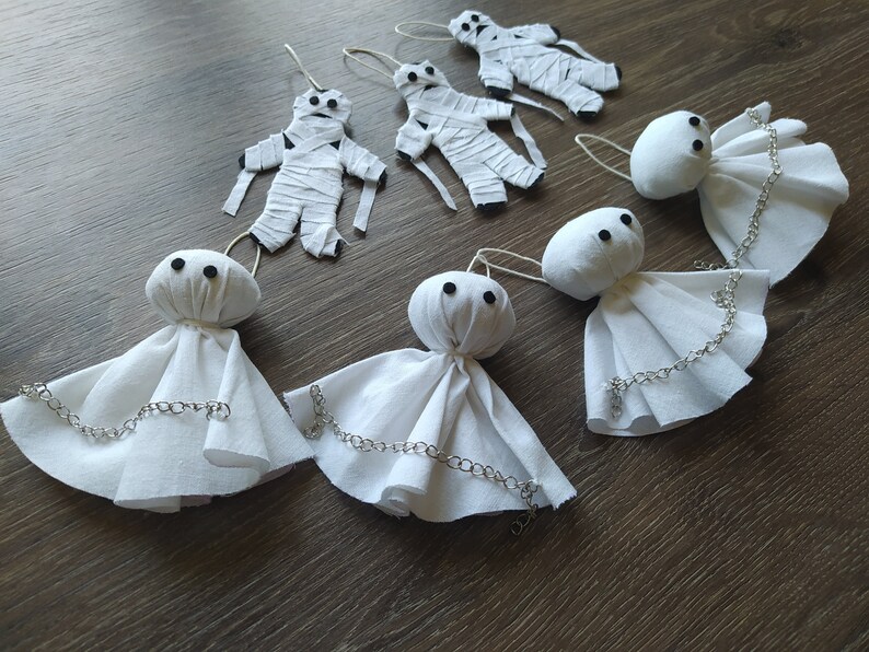 May include: Three white fabric ghost ornaments with black bead eyes and silver-toned chains draped across their skirts, alongside three white fabric mummy ornaments wrapped in strips with black bead eyes, all featuring small hanging loops.