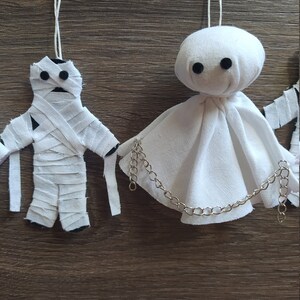 May include: Two hanging ornaments made of white fabric. One is a mummy figure wrapped in strips of cloth with black circular eyes. The other is a ghost figure with a rounded head, a draped body, black circular eyes, and a silver metal chain.
