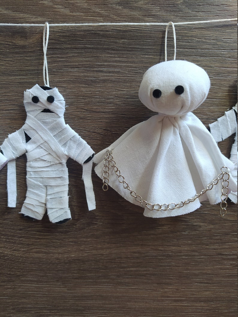 May include: Two hanging ornaments made of white fabric. One is a mummy figure wrapped in strips of cloth with black circular eyes. The other is a ghost figure with a rounded head, a draped body, black circular eyes, and a silver metal chain.