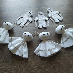 May include: A collection of seven handmade ornaments consisting of three white fabric mummy figures wrapped in strips and four white ghost figures with round heads, black bead eyes, and silver-toned metal chains draped across their fabric bodies.